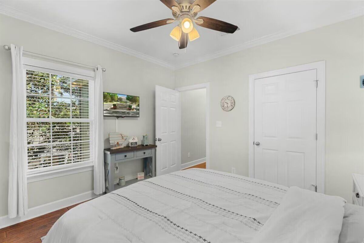 A bedroom with a bed, a ceiling fan, a window with blinds, a desk, and a door.