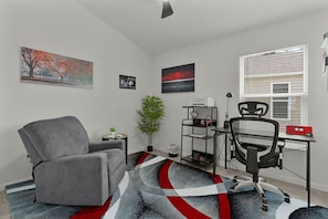 A modern office space with a grey armchair, a desk with a computer, a bookshelf, and a window with blinds. - Bright & Pet-Friendly Cottage Home in the Village of Well Point (Sumterville)