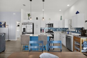 A modern kitchen with white cabinetry, a stainless steel refrigerator, a built-in oven, and a microwave. There are blue chairs and a small dining table. - Bright & Pet-Friendly Cottage Home in the Village of Well Point (Sumterville)