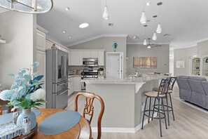 A modern kitchen with stainless steel appliances, a central island with bar stools, and a dining area with a wooden table and chairs. - 3/2 Designer in Village of Mallory Square Dog friendly w/ Golf cart (The Villages)