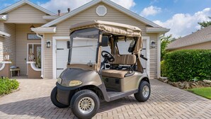 A golf cart parked in front of a house with a garage, a driveway, and a hedge. - 3/2 Designer in Village of Mallory Square Dog friendly w/ Golf cart (The Villages)