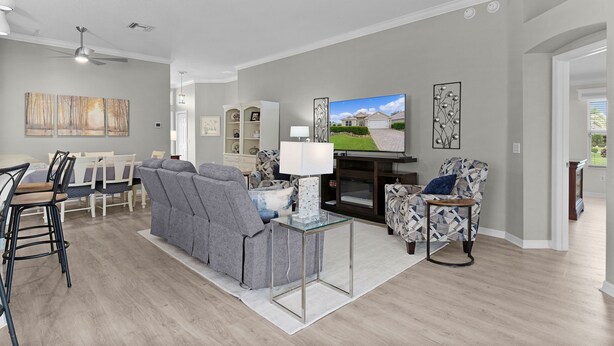A modern living room with a flat-screen TV, grey sofas, a glass coffee table, and a dining area with chairs and a table. - 3/2 Designer in Village of Mallory Square Dog friendly w/ Golf cart (The Villages)