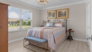 A bedroom with a bed, bench, bedside table, lamp, and two framed pictures on the wall. - 3/2 Designer in Village of Mallory Square Dog friendly w/ Golf cart (The Villages)