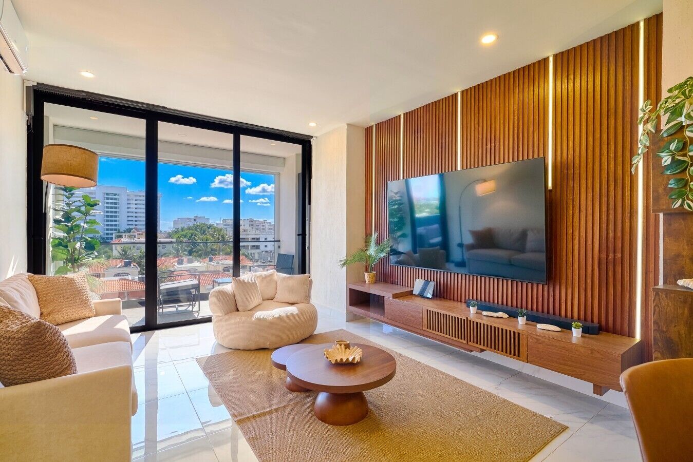 A modern living room with a large flat-screen TV, a sofa, a coffee table, and a balcony with a city view.