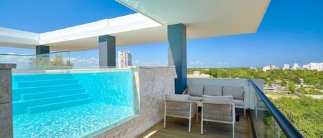 A modern balcony with a built-in swimming pool, a seating area with a sofa and chairs, and a clear view of the surrounding landscape.