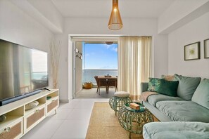 A modern living room with a flat-screen TV, a sofa, a coffee table, and a view of the ocean through a sliding door. - FrontBeach Apt Pool AC Juan Dolio Beach Aquarella (Playa Juan Dolio)