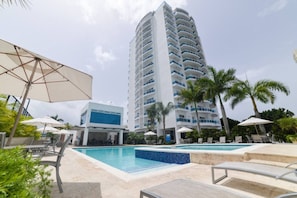 A hotel with a pool area, palm trees, and a tall building in the background. - Tropical BeachSide Apartment 4PAX AC La Velas (Playa Juan Dolio)