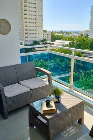 A balcony with a wicker sofa and a small table, a clear view of a green landscape, and a tall building in the background. - Tropical BeachSide Apartment 4PAX AC La Velas (Playa Juan Dolio)