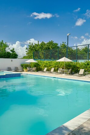 Ein Swimmingpool mit Liegestühlen und Sonnenschirmen, umgeben von einem Zaun und viel Grün.
