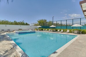 Ein rechteckiger Swimmingpool mit gefliestem Rand, umgeben von Liegestühlen und Sonnenschirmen, begrenzt durch einen Zaun.