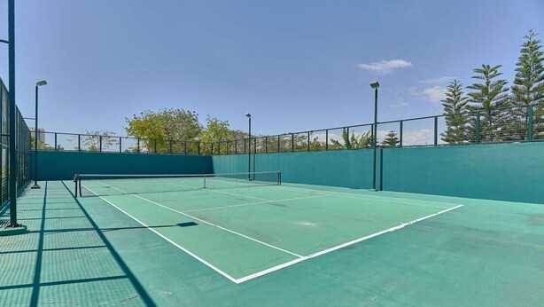 En udendørs tennisbane med net, omgivet af et grønt hegn og træer.