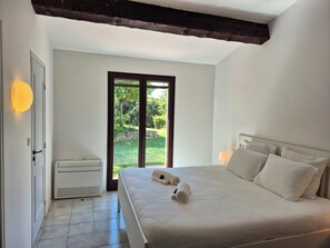 Quarto com cama, mesa de cabeceira, abajur e vista para um jardim através de uma porta de vidro.