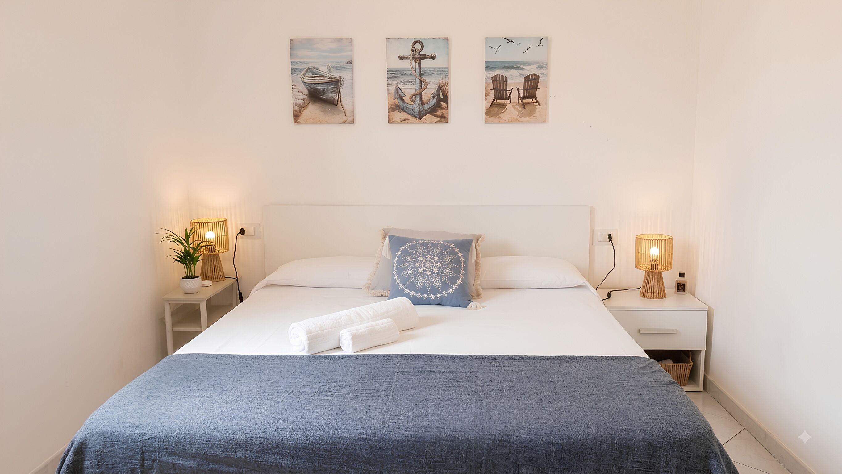 Un lit bien fait, avec une couverture bleue et des oreillers, deux lampes de chevet et trois tableaux accrochés au mur représentant des scènes de plage.