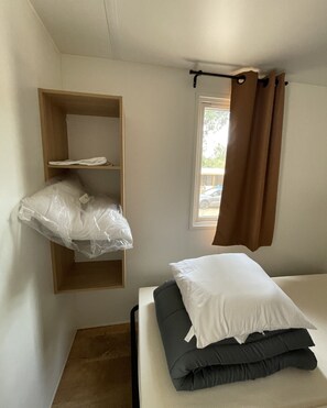 A small, single-bed room with a wooden shelf, a window with brown curtains, and plastic bags hanging from the shelf. - New bungalow at a 3-star campground—right on the water (Porto-Vecchio)