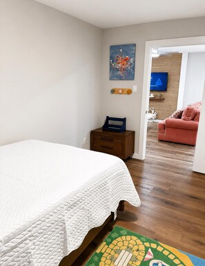 A bedroom with a bed, a wooden nightstand, a blue chair, a red sofa, and a television in the adjacent living room. - Home: 3 Minutes to Sylvan Beach with a Hot Tub and Office Space (La Porte)
