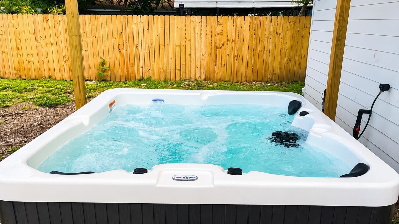 A hot tub in a backyard with a wooden fence and a white house in the background.