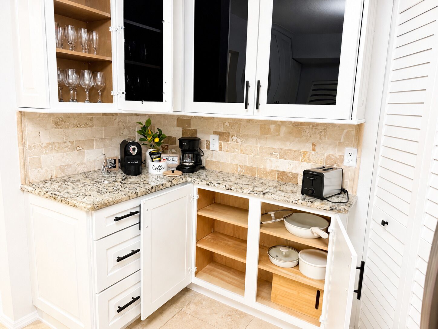 Una cocina con armarios blancos, cafetera, tostadora y estante con cristalería.