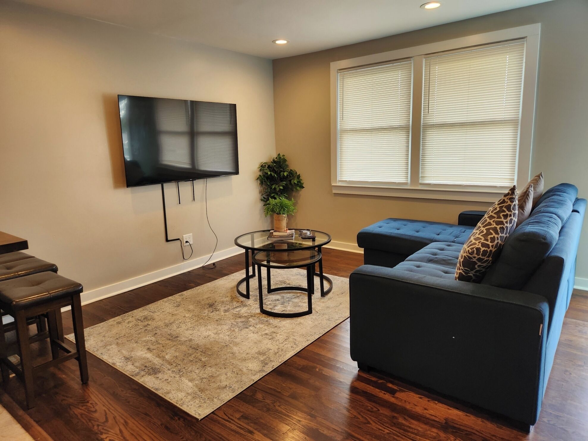 A living room with a flat-screen TV mounted on the wall, a round glass coffee table, a dark blue sofa with patterned cushions, and a wooden dining table with bar stools.
