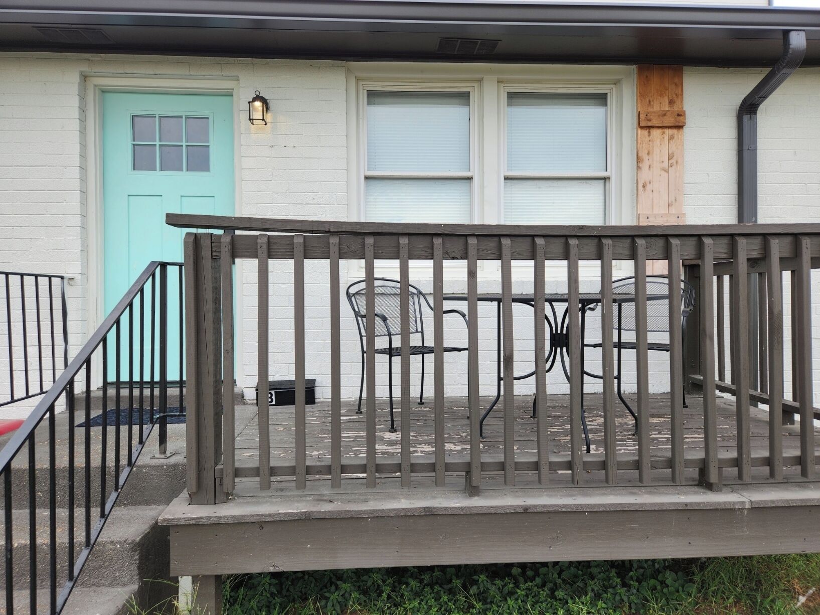 Une terrasse en bois avec une balustrade, une porte turquoise et une fenêtre avec des stores.