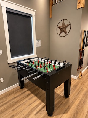 A foosball table in a room with a window, a star decoration on the wall, and a rack of sports equipment. - Texas sized family fun with basketball, pickleball, gym, arcade, billiards etc.. (Anna)
