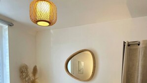 Una cama bien hecha con almohadas y una manta, una lámpara de noche, un espejo en la pared y una ventana con cortinas.