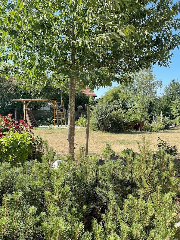 Un jardín con un parque infantil de madera, un árbol y varios arbustos.