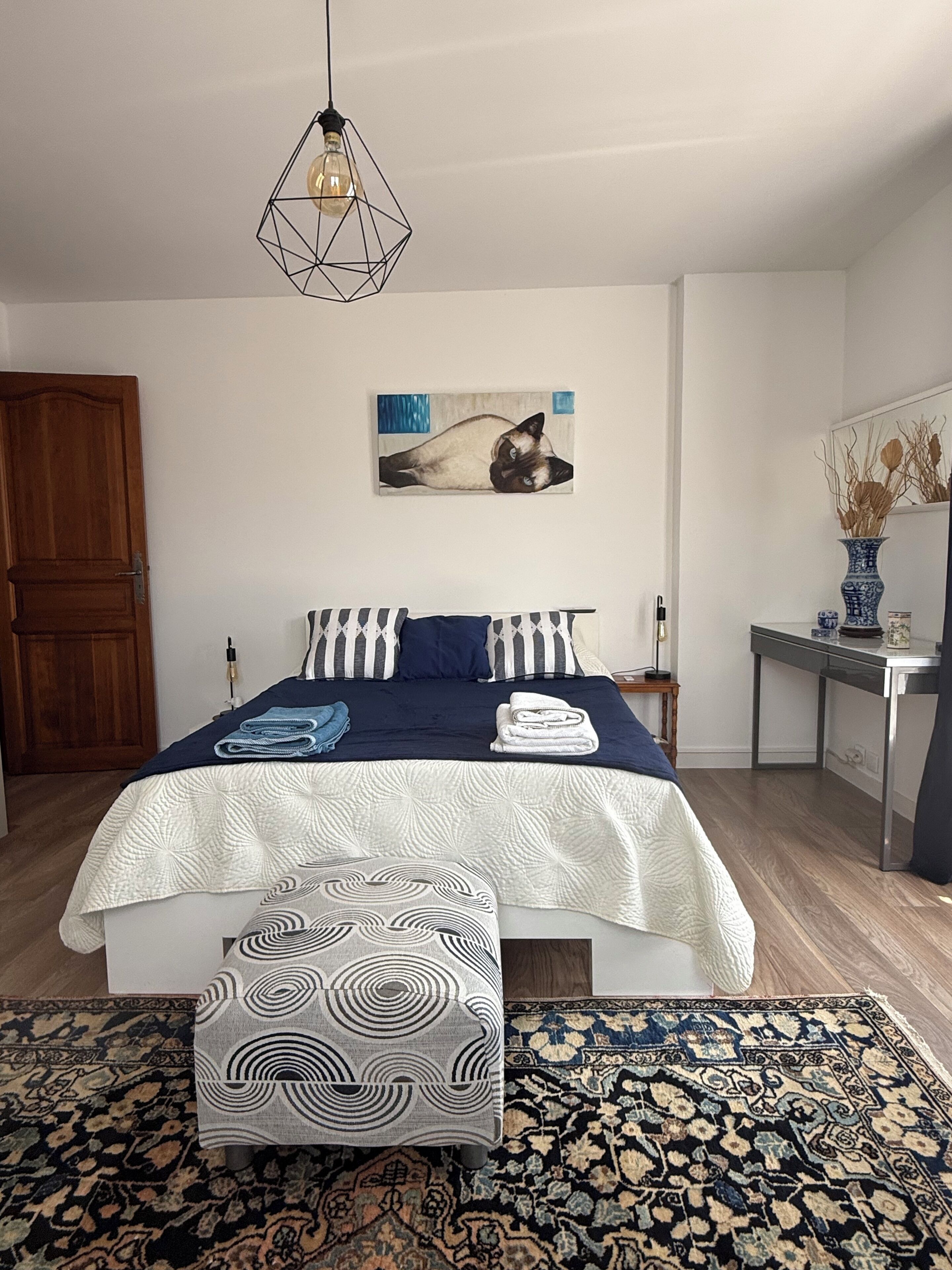 Un dormitorio con cama, mesita de noche, silla, alfombra, cuadro y puerta de madera.
