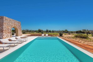 Una piscina rectangular amb aigua blava i cristal·lina, envoltada de gandules blanques i una estructura de pedra amb un arc.