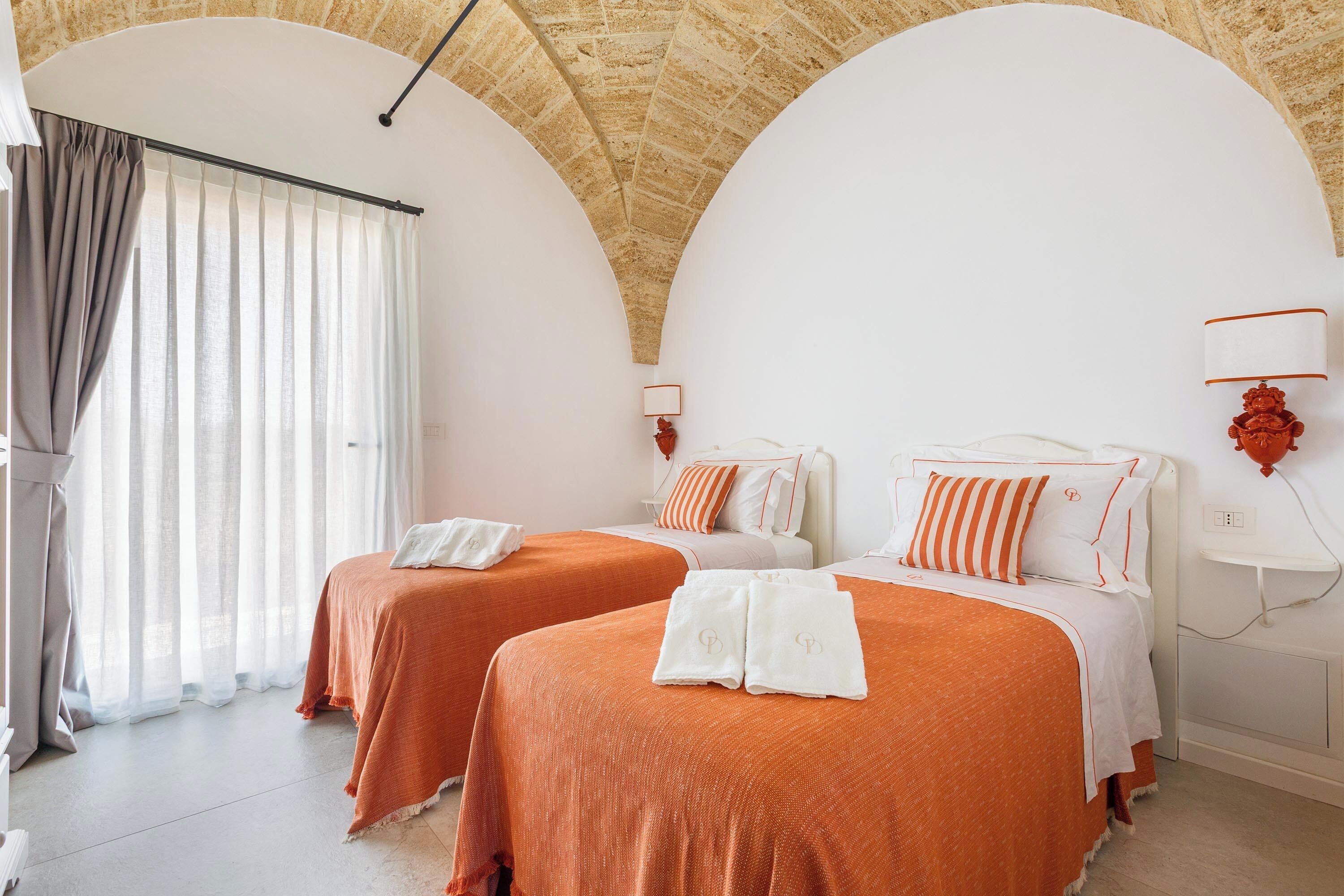 Een hotelkamer met twee bedden, een groot raam met gordijnen, een wandlamp en een betegeld plafond.