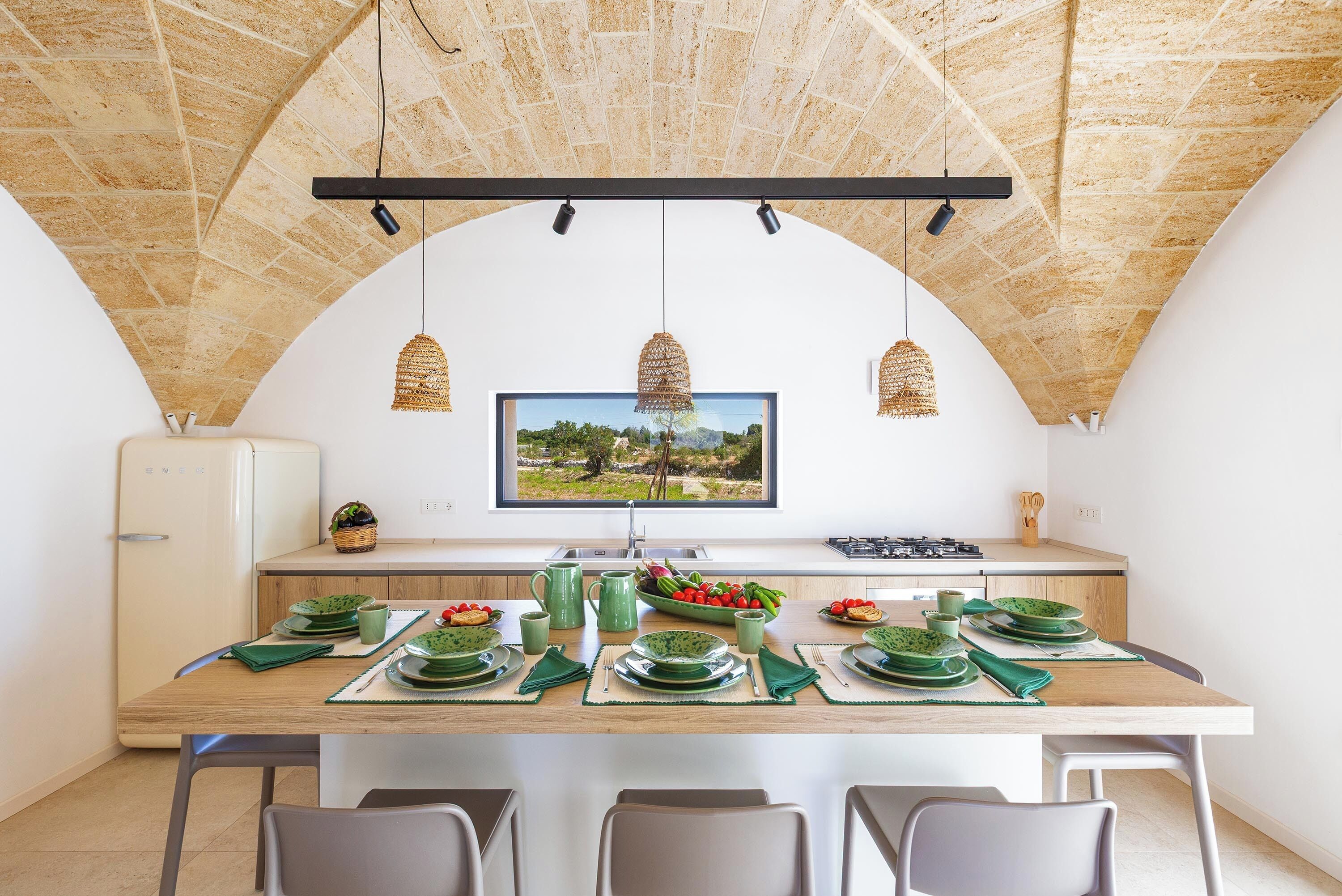 A dining area with a long table set for four, a television mounted on the wall, and a view of a landscape through the window.