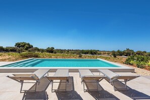 A rectangular swimming pool with clear blue water, surrounded by a low stone wall. In the foreground, there are three sun loungers aligned parallel to the pool. The background features a clear sky and a landscape with trees and shrubs. - Onirica Dimora by BarbarHouse (Salve)