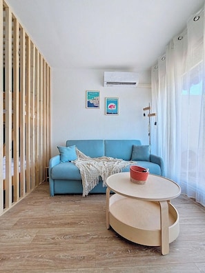 A room with a blue sofa, a round wooden table, and two wall-mounted air conditioning units. - Jardins du Port D3073 – Studio clim, terrasse, parking, Wi-Fi, piscine (Le Grau-du-Roi)