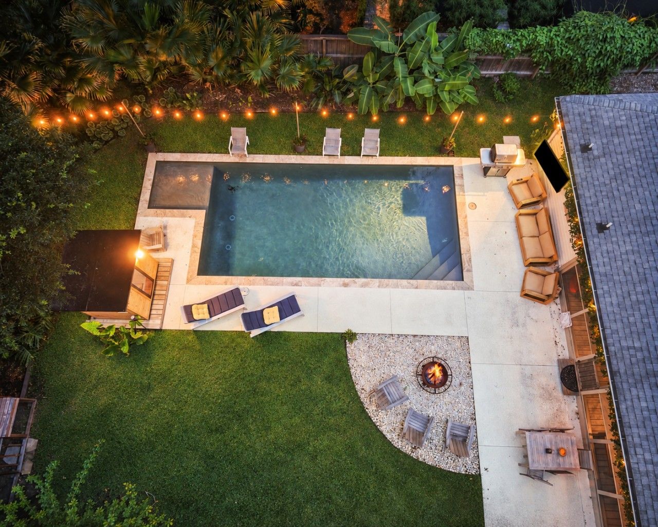 An aerial view of a backyard with a swimming pool, lounge chairs, a fire pit, and string lights.