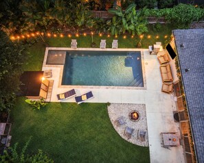 Ein Blick von oben auf einen Garten mit Schwimmbad, Liegestühlen, Feuerstelle und Lichterketten.