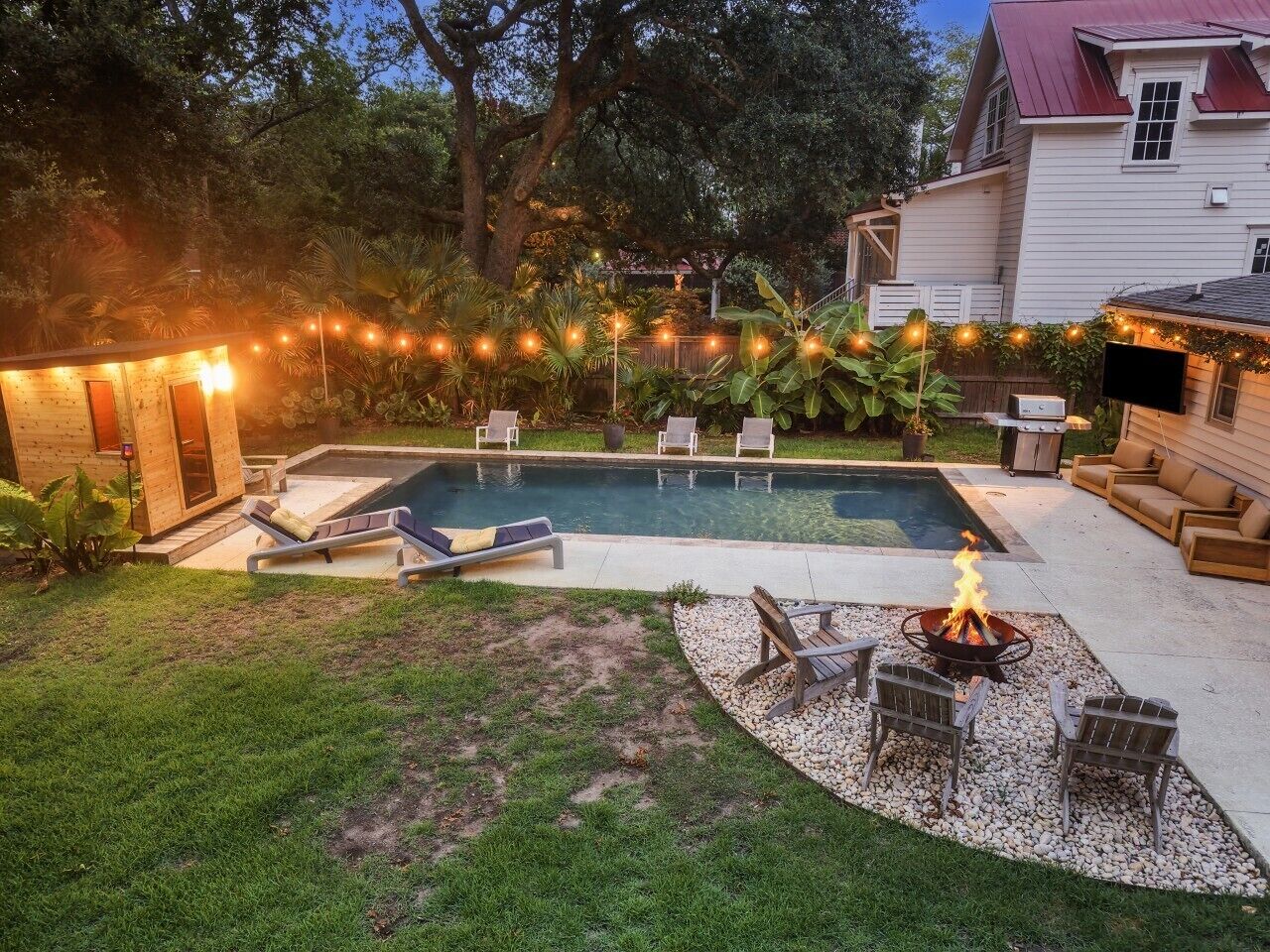 A backyard with a swimming pool, lounge chairs, a fire pit, and string lights.