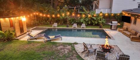 A backyard with a swimming pool, lounge chairs, a fire pit, and string lights.