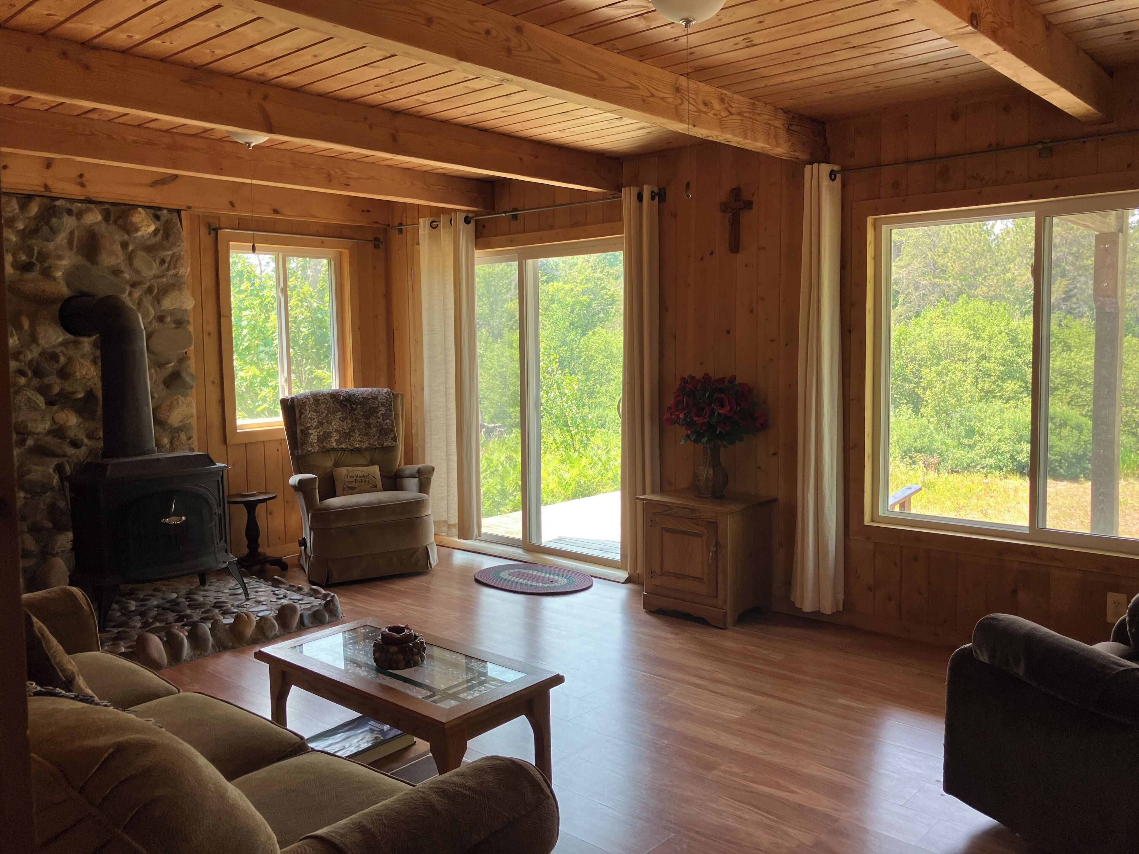 A cozy cabin interior with a stone wall, a wood-burning stove, a comfortable armchair, a sofa, a coffee table, and large windows offering a view of the outdoors.