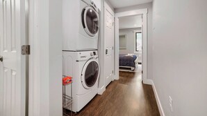 A laundry room with stacked washers and dryers, a wooden floor, and a hallway leading to a bedroom with a bed and window. - NEW - Charming 2-Bedroom Condo Near Shuttle Stops and Downtown (Breckenridge)