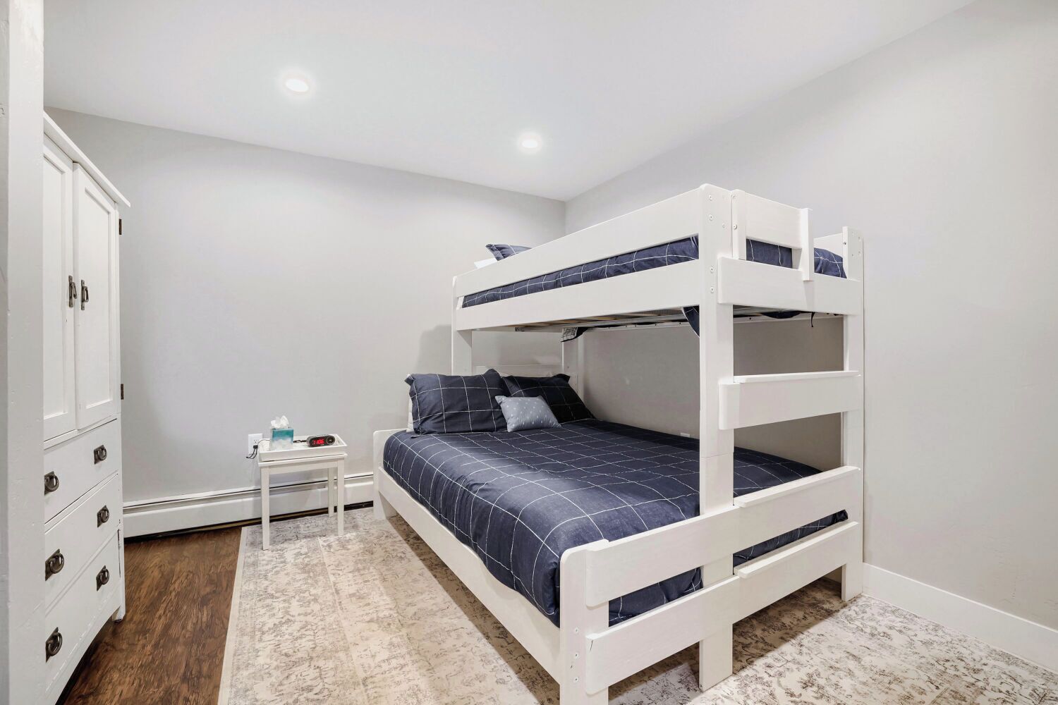 A bunk bed room with a white wardrobe, a small bedside table with a clock, and a patterned rug.