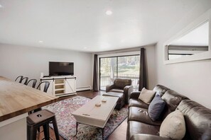 A modern living room with a dining area, a sofa set, a flat-screen TV, and a large window offering a view of the outdoors. - NEW - Charming 2-Bedroom Condo Near Shuttle Stops and Downtown (Breckenridge)
