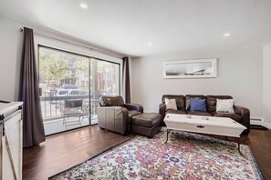 A modern living room with a sofa, ottoman, and coffee table, a large window with curtains, and a patterned rug. - NEW - Charming 2-Bedroom Condo Near Shuttle Stops and Downtown (Breckenridge)