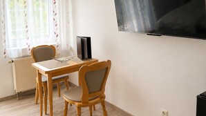 A room with a dining table, two chairs, a radiator, a TV on the wall, and a window with curtains.