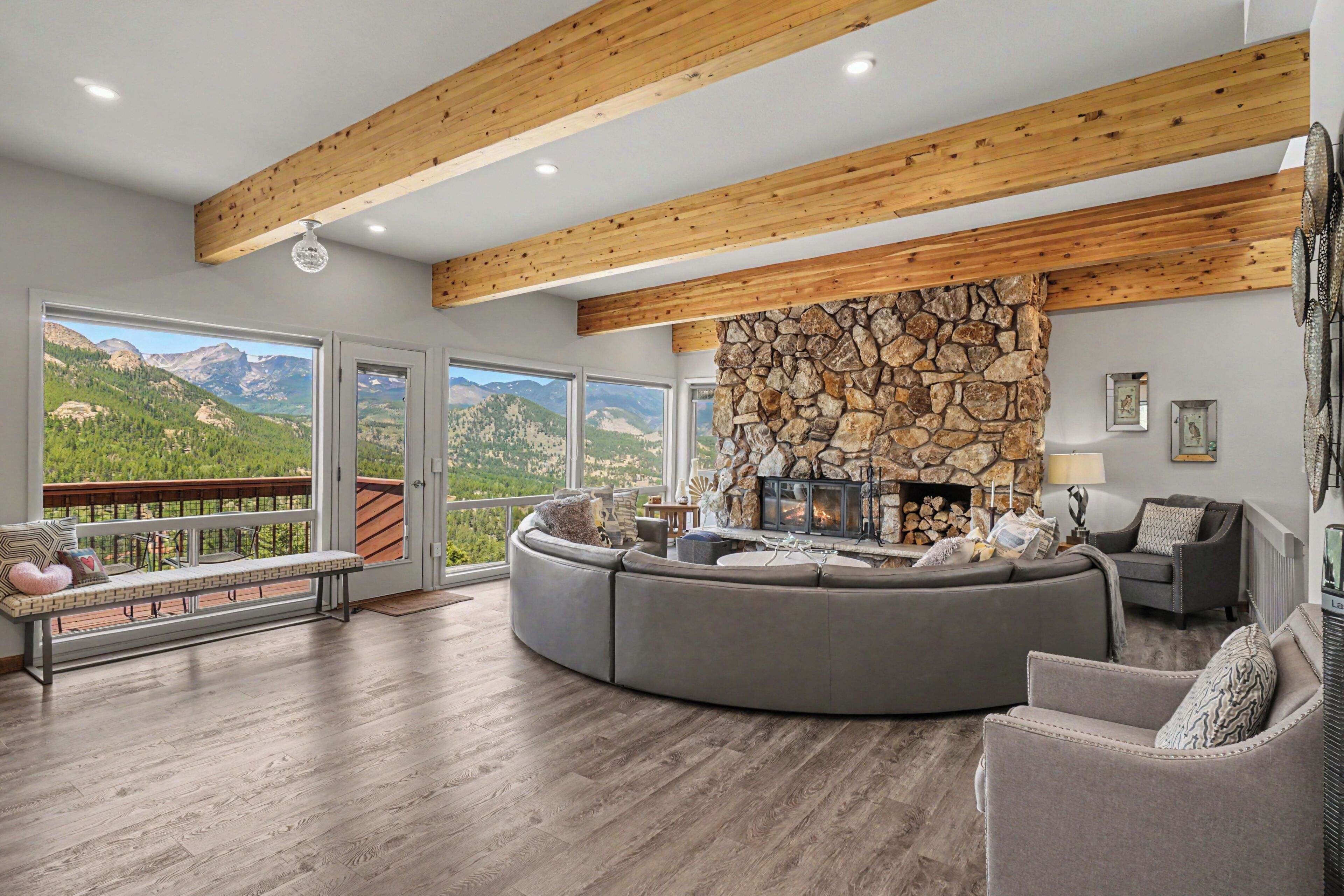 A spacious living room with a stone fireplace, a curved grey sofa, and a view of mountains through large windows.