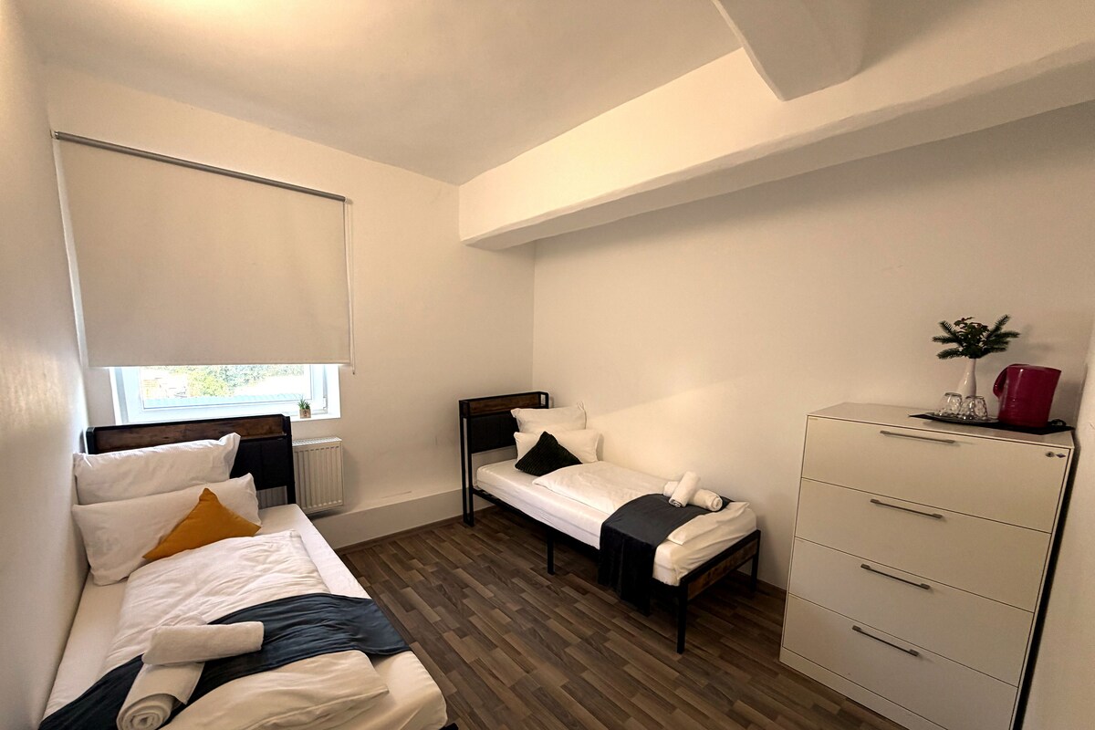 A room with two beds, a dresser, a radiator, and a window with blinds.