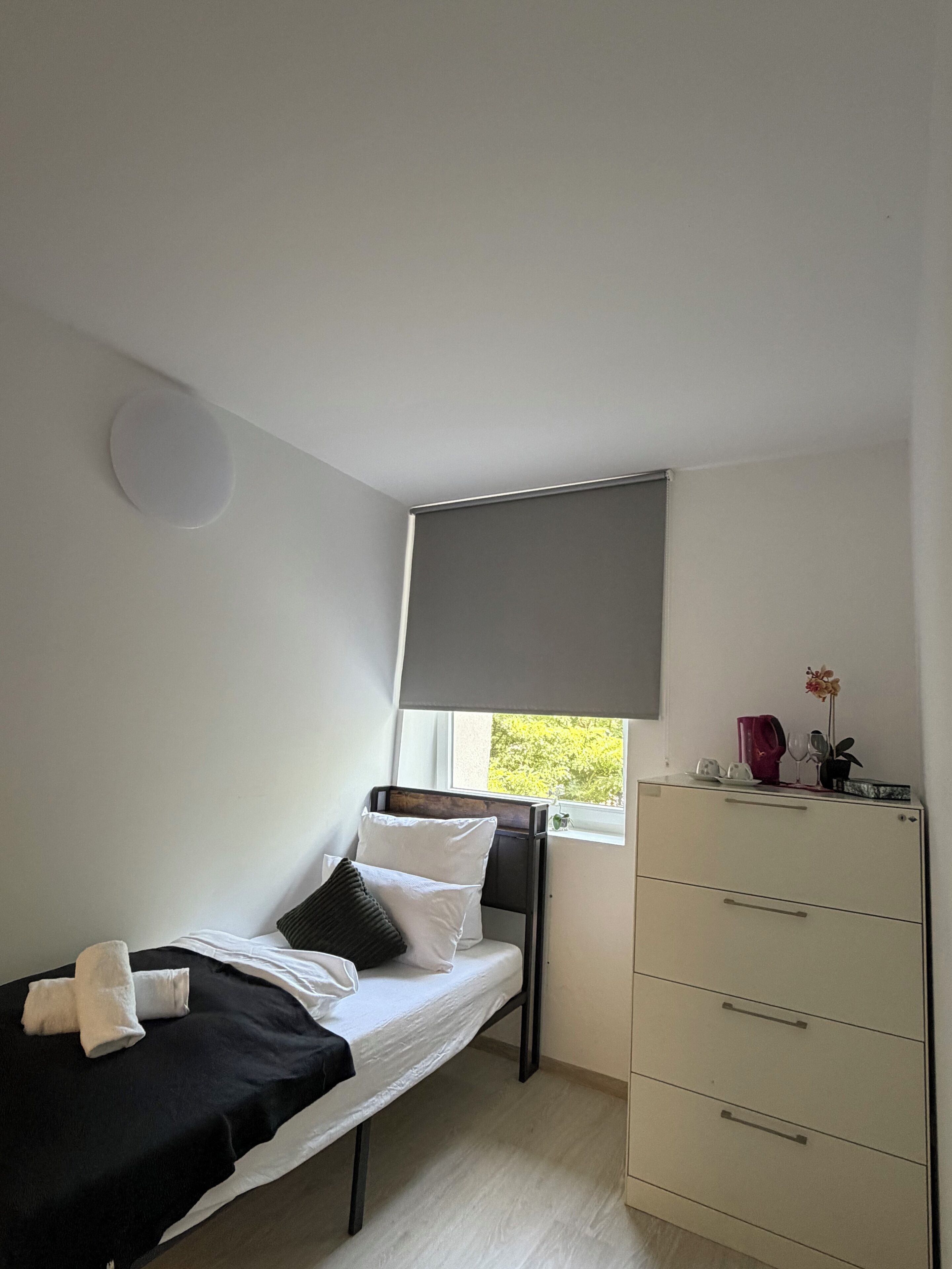 Ein kleines Zimmer mit einem Einzelbett, einem Fenster, einem Schreibtisch mit Schubladen und einer Heizung.