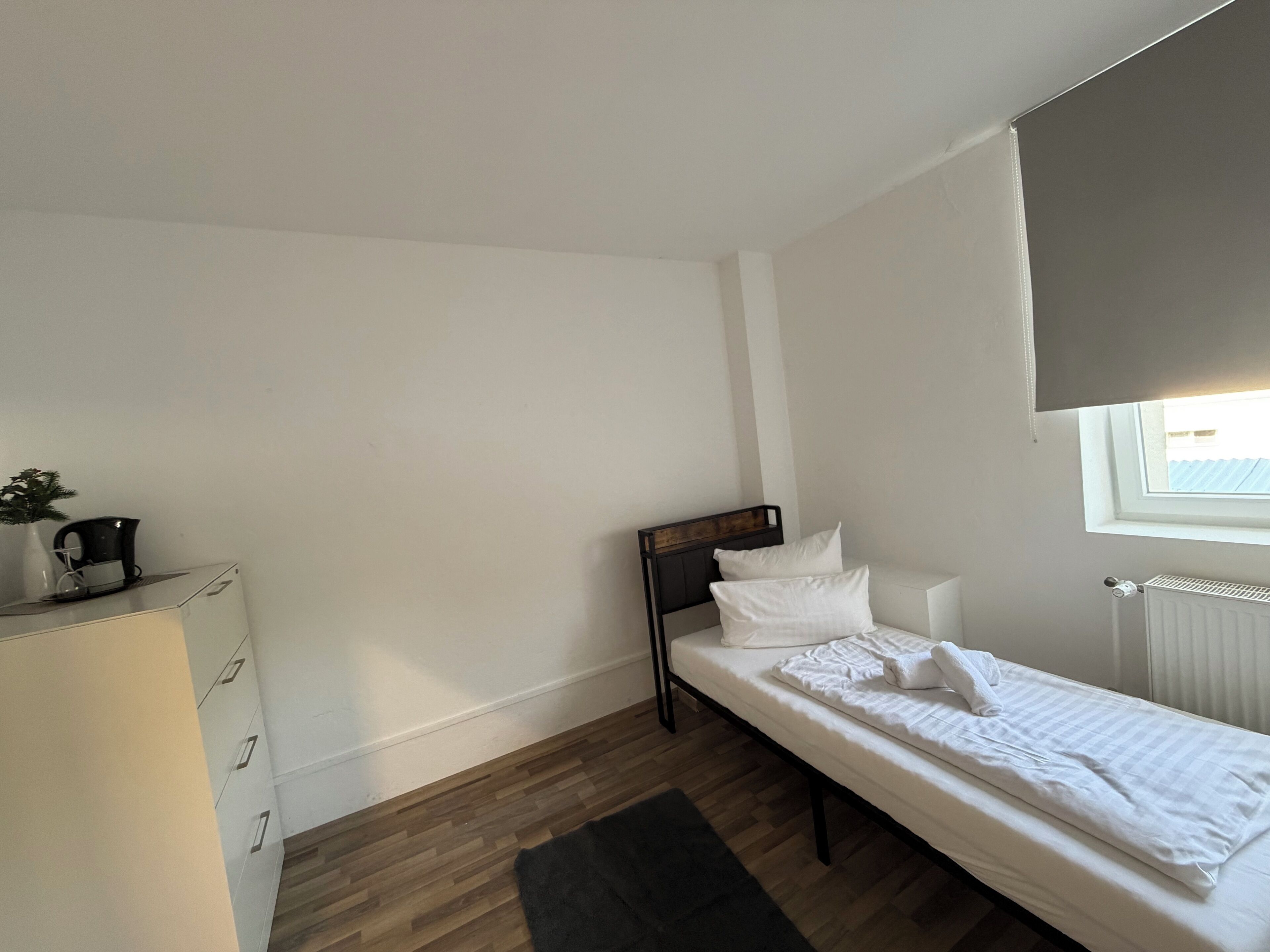 A small, tidy room with a single bed, a dresser, a radiator, and a window with a blind.
