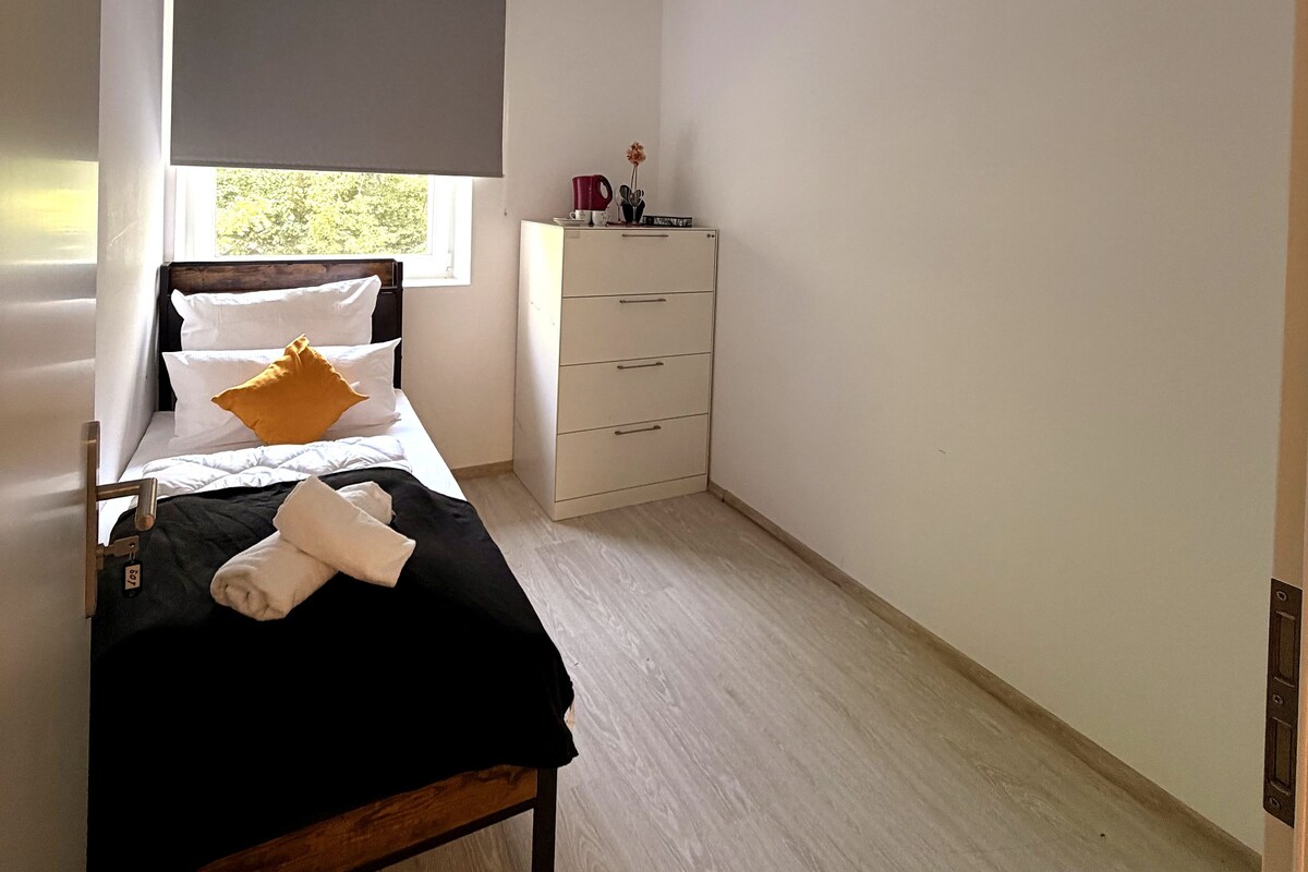 A bedroom with a bed, a bedside table, a dresser, and a window with a blind.