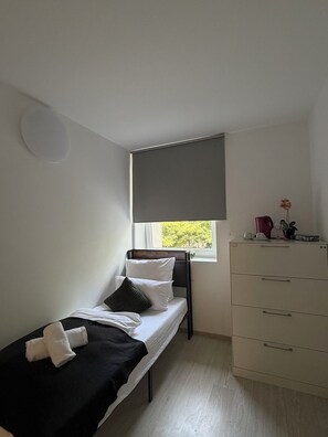 A small, single-bed room with a window, a bedside table, and a dresser.