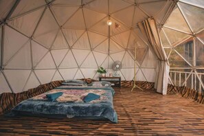 A tent with a geometric ceiling structure, a bed with a patterned bedspread, and a small table with a plant.