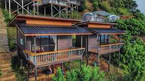A modern two-story house with a balcony, surrounded by lush greenery.
